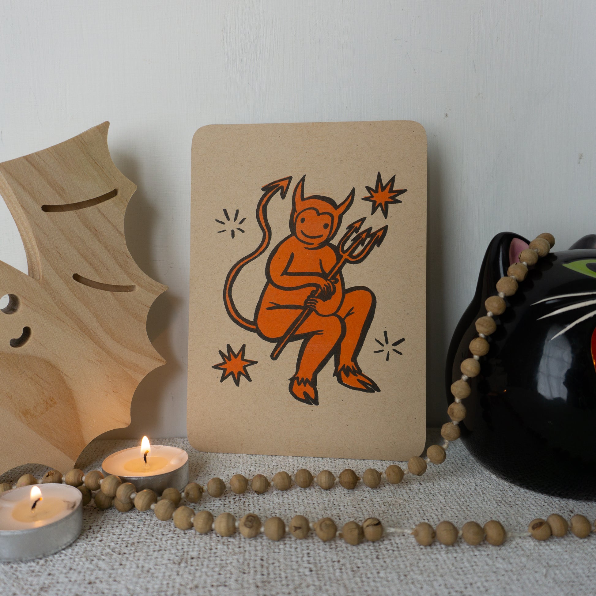 Orange and black illustration of a cartoon devil on kraft color paper with rounded corners.
