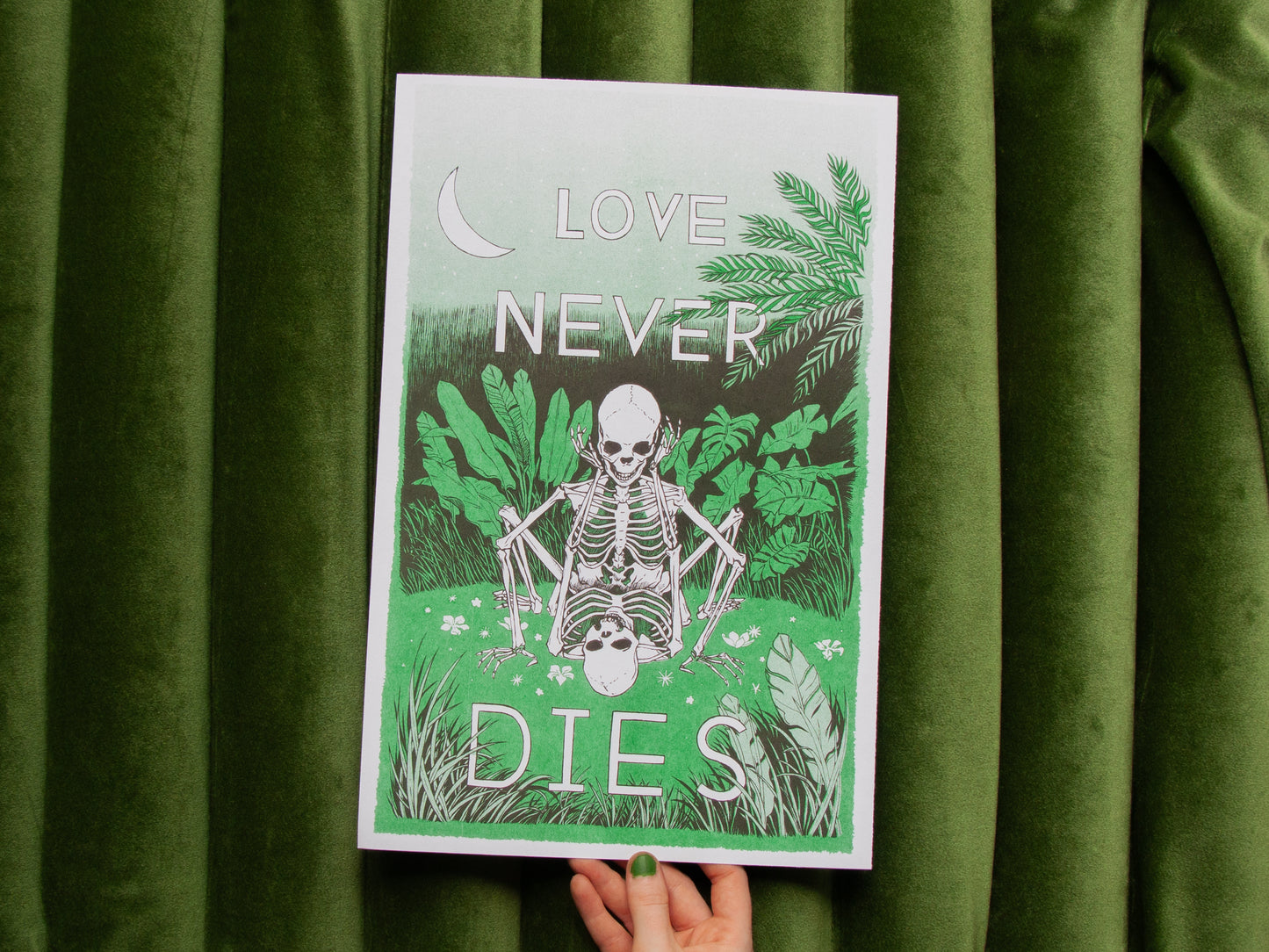 Two skeletons in love are doing it in a field of green. The print is held by hand over green fabric, and is 10 inches by 16 inches. It is an illustration printed on a risograph in green and black inks.