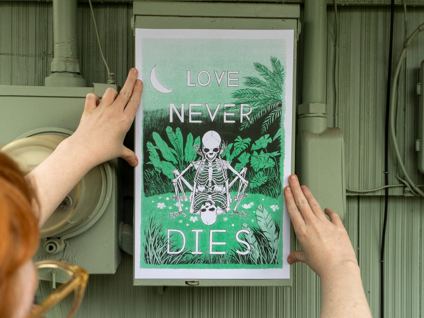A person holding a risograph print with both hands against an electrical box. The print has two skeletons in love doing it in a field of green.