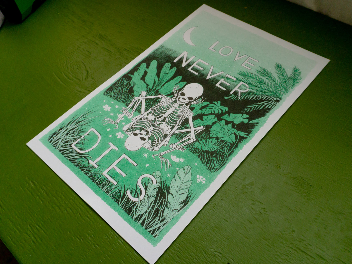 A risograph print laying on a green, painted table. It has two skeletons in love doing it in a field of green.