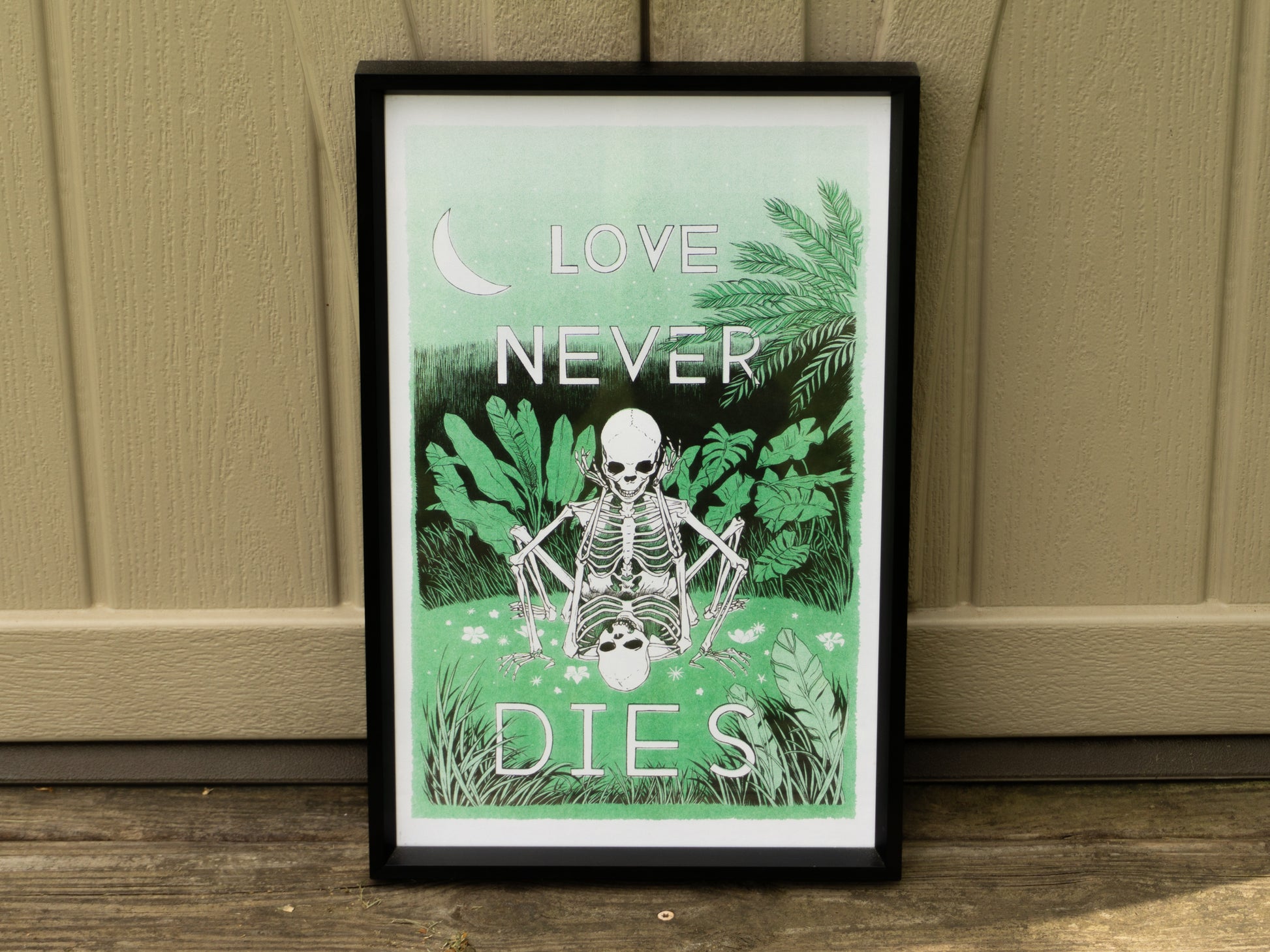 A risograph print framed in a black picture frame outside. The artwork has two skeletons in love doing it in a field of green.