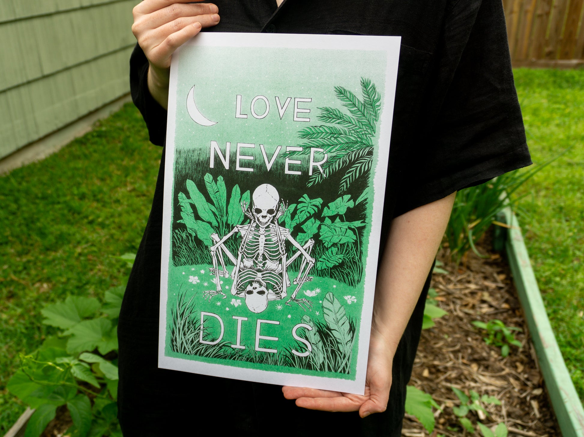 A person wearing black holds a riso print, 10 inches x 16 inches, in front of a garden. The print has two skeletons in love doing it in a field of green.