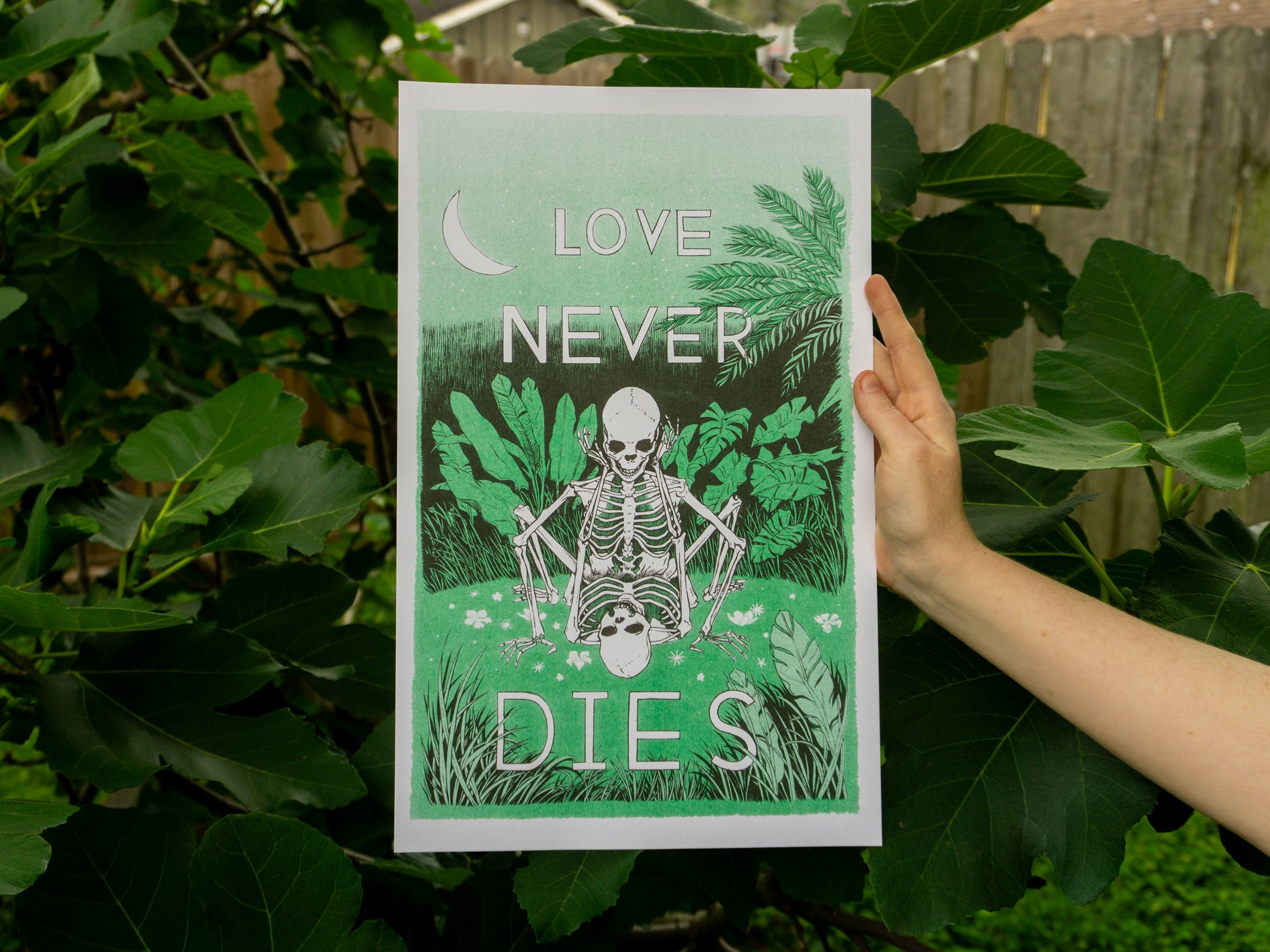 A hand holds a 10 inches x 16 inches art print in front of a fig tree. In the print, two skeletons in love are doing it in a field of green.