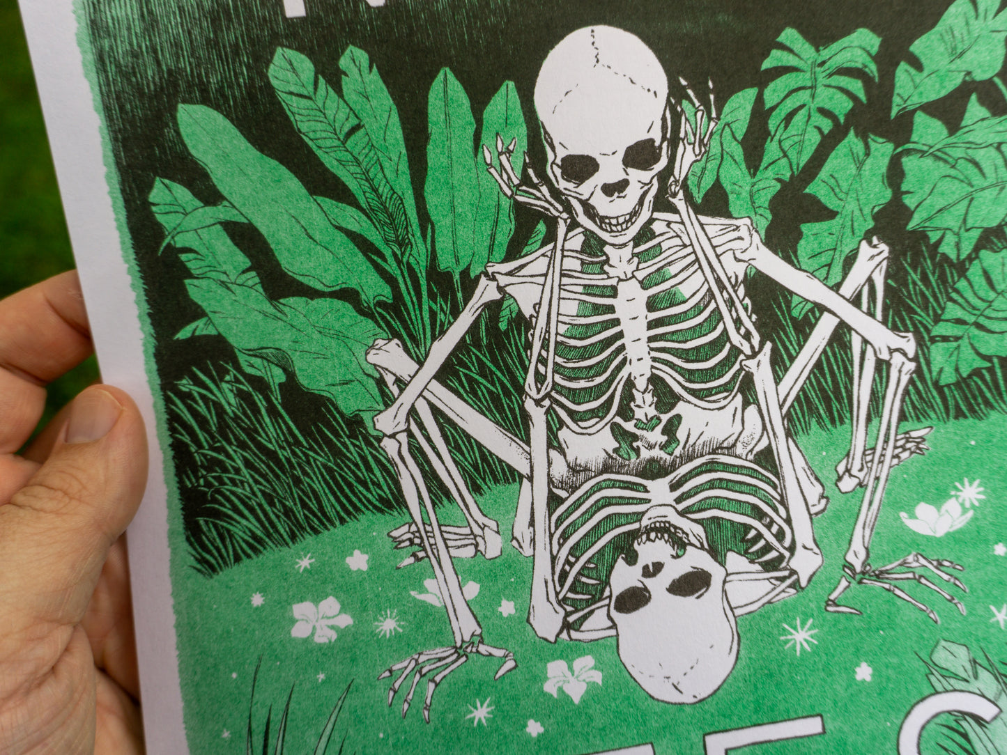 Close up image of an art print on paper held in one hand. The artwork has two skeletons doing it behind some bushes.