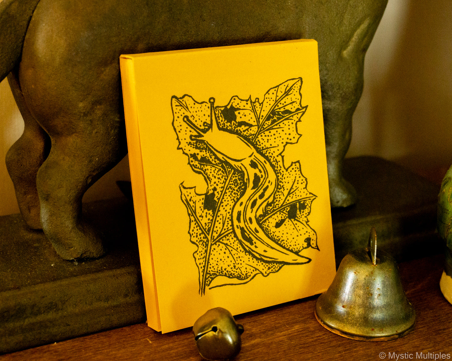 Yellow notebook with a banana slug and leaf design on a wooden surface with bells and a statue.