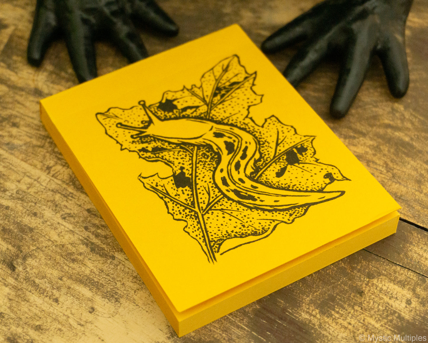 Yellow notepad with black leaf design and slug on a wooden surface with black frog feet.