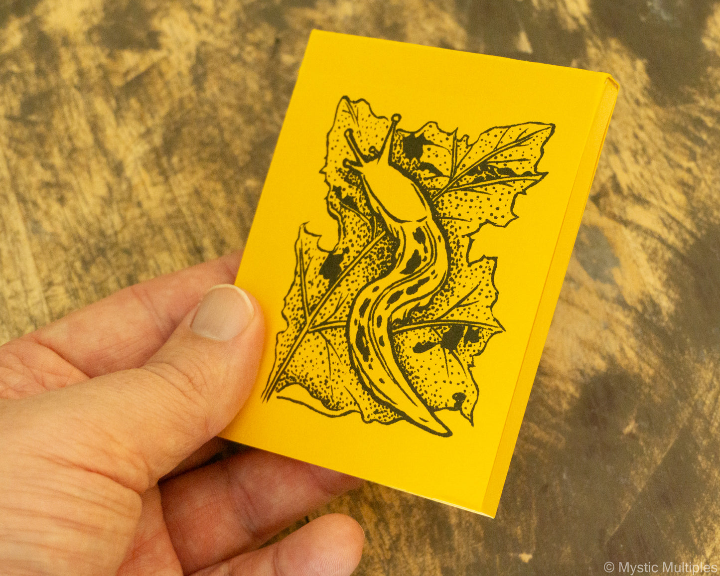Yellow notebook with a black illustration of a leaf and a small slug held by a hand on a wooden surface.