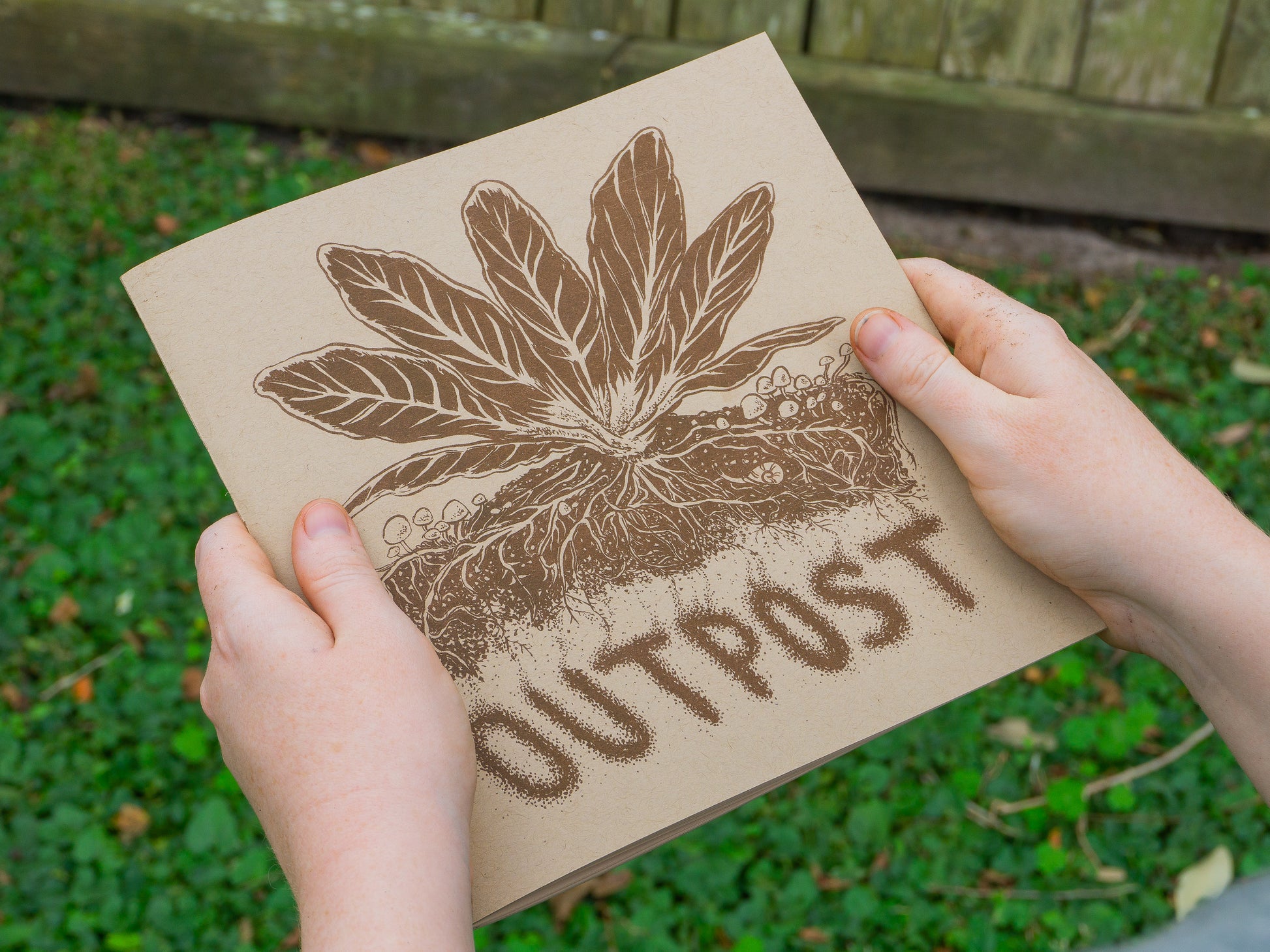 Cover of the zine with a mustard plant growing among mushrooms with an underground view of the root structure. Hands demonstrate the scale of 8 by 8.