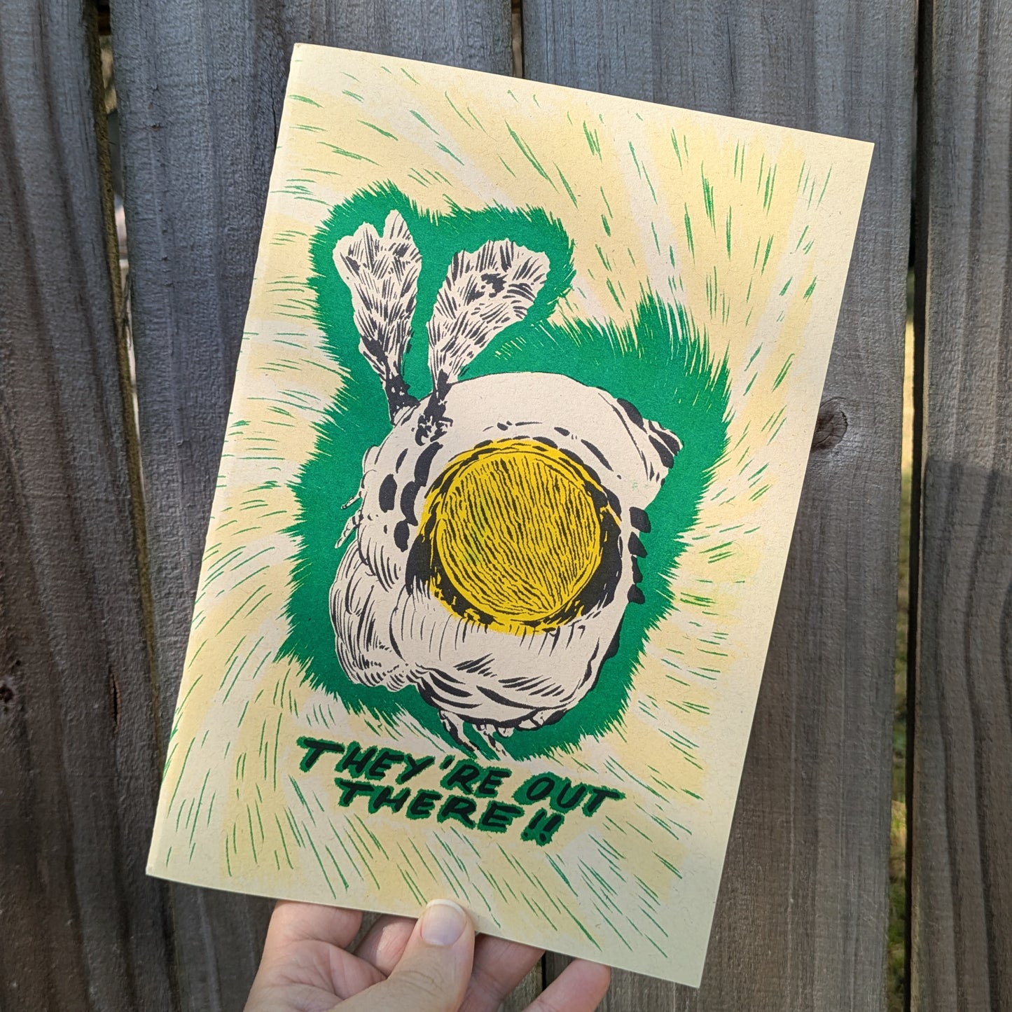 Hand holding a zine with a graphic of a wasp and text 'They're Out There!' against a wooden background