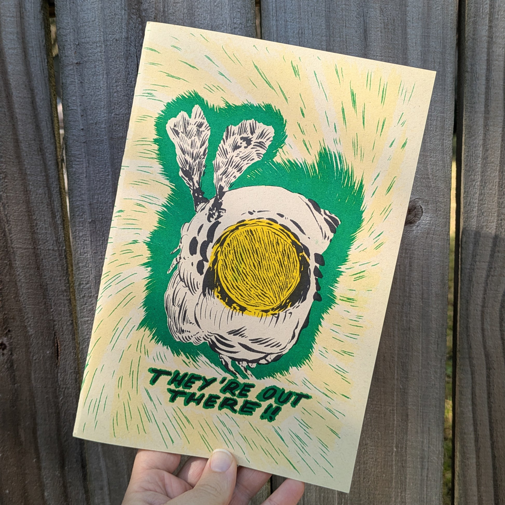 Hand holding a zine with a graphic of a wasp and text 'They're Out There!' against a wooden background